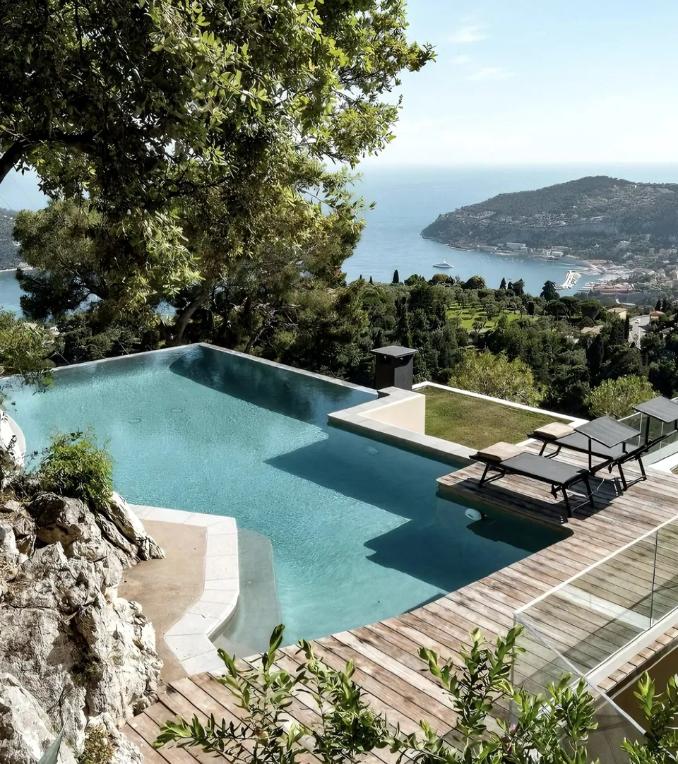 Piscine à débordement et terrasse en bois surplombant une baie.